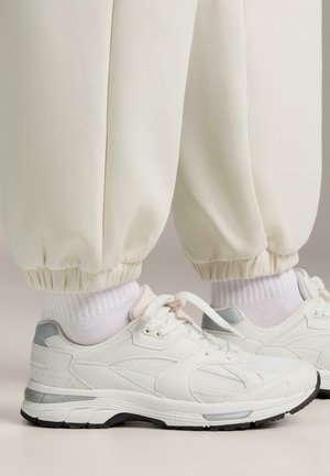 Scarpe da ginnastica bianche con tomaia in mesh e sintetico, dettagli grigi, design con lacci, abbinate a calze bianche a coste e pantaloni jogger color crema.