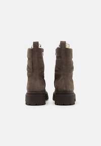 Brūni zamšādas potītes zābaki ar teksturētu apdari, ar masīvu tumšu zoli un vilkšanas cilpu ērtai uzvilkšanai, parādot šuvju detaļas.