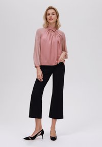 Blusa in chiffon rosa blush con scollatura plissettata e maniche lunghe trasparenti, abbinata a pantaloni neri a vita alta e tacchi neri a punta.
