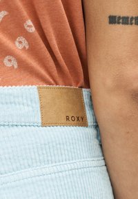 Κοντινό πλάνο ενός ατόμου που φοράει ανοιχτό γαλάζιο παντελόνι κοτλέ με καφέ ετικέτα «ROXY» και πουκάμισο σκουροκόκκινο χρώμα, με μερικώς ορατό τατουάζ.