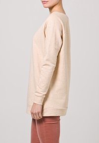 Béžová mikina s dlouhým rukávem se skvrnitou texturou, kulatým výstřihem a bočními rozparky. Kombinována s těsně přiléhavými světle růžovými kalhotami.