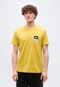 Jeune homme aux cheveux bouclés portant un T-shirt jaune avec un écusson montagne noir et blanc marqué "BOSS" sur la poitrine gauche.