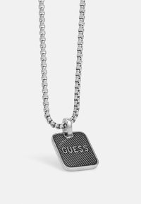 Zilveren ketting met een zwart rechthoekig hangertje met een gestructureerd oppervlak en het woord "GUESS" erop geperst.