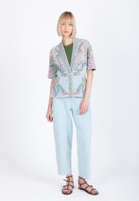 Naine blondide bobidega, kandes pastelltoonides mustriga lühikeste varrukatega kardigani, rohelist toppi, helesiniseid pükse ja pruune gladiaatorstiilis sandaale, seistes.