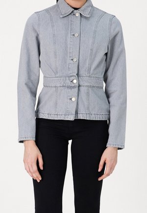Femme portant une veste en jean gris clair boutonnée avec une couture à la taille et un pantalon noir, montrée des épaules aux hanches.
