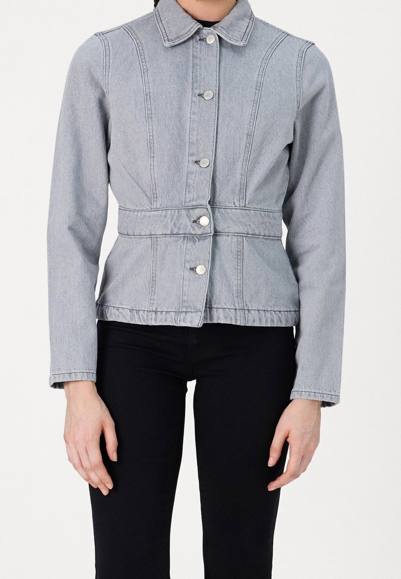 Femme portant une veste en jean gris clair boutonnée avec une couture à la taille et un pantalon noir, montrée des épaules aux hanches.