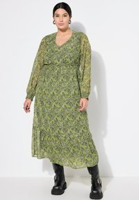 Abito floreale verde con maniche lunghe trasparenti, scollatura a V, vita arricciata e gonna a strati; abbinato a stivali militari neri.