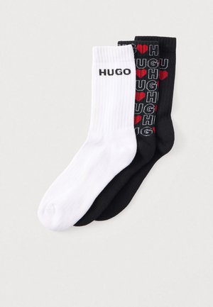 Pack de dois pares de meias em preto e branco. A meia branca apresenta o texto "HUGO"; a meia preta tem um padrão repetido "HUGH" com corações vermelhos. Textura canelada.