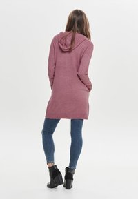 Langer, kapuzenpullover aus weichem, rosa Strickstoff, mit geripptem Saum und Bündchen, kombiniert mit engen blauen Jeans und schwarzen, hohen Stiefeletten.