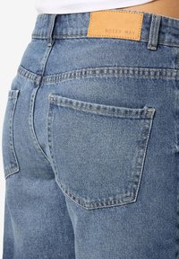 Zilas denima bikses ar nolietotu apdari, kurām ir brūns ādas jostas plāksteris ar uzrakstu "NOISY MAY" un divas aizmugures kabatas.