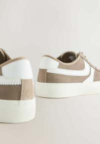 Brune ruskinds sneakers med hvide læderdetaljer, rund form, flad hvid sål og struktureret syning langs siderne.