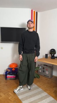 Must must särk, lahtised oliivrohelised laiad püksid, hallid ja valged tossud. Seisab triibulisel vaibal modernse sisustusega ruumis.