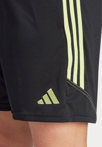 Pantaloni sportivi neri con strisce verde lime e un logo. Presentano una texture liscia e tasche laterali. Il design include cuciture angolari.