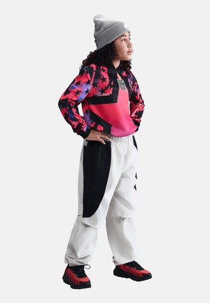 Młoda osoba w szarej czapce, żywym czerwono-czarnym bluzie z kapturem z wzorem gwiazd i emblematem USA, białych i czarnych spodniach oraz czerwono-czarnych trampkach, z rękami na biodrach.