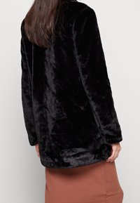 Mujer vestida con una chaqueta de piel sintética negra y una falda de canalé marrón, vista desde atrás contra un fondo claro liso.