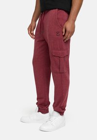 Burgunderfarbene, lässige Jogginghosen aus weichem Stoff mit einer seitlichen Cargo-Tasche, Kordelzugbund und schwarzen Logo-Details. Kombiniert mit weißen Sneakers.