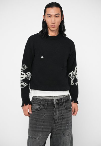 Pull en tricot noir avec un col rond, des détails usés et des motifs de crane blancs sur les manches. Associé à un jean gris.