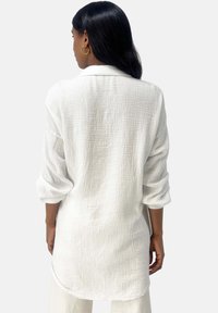 Chemise blanche texturée à manches longues avec des poignets retroussés et un ourlet arrondi. Tissu léger avec un motif froissé. Vue arrière montrée.