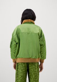 Grön bomberjacka med en beige fuskpälskrage, med en blank finish, ribbat nederkant och en avslappnad passform. Mönstrade gröna byxor.