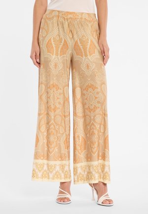 Wijde beige broek met oranje paisley-patronen, gedragen met witte sandalen met hak en bandjes en een wit topje in de broek gestopt.