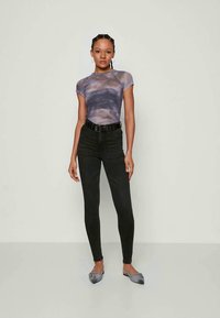 Νεαρή γυναίκα με πλεξούδες, που φοράει ένα λιτό μπλουζάκι τύπου tie-dye σε μωβ χρώμα, μαύρα skinny τζιν με ψηλή μέση, ζώνη και γκρι μυτερές γόβες.