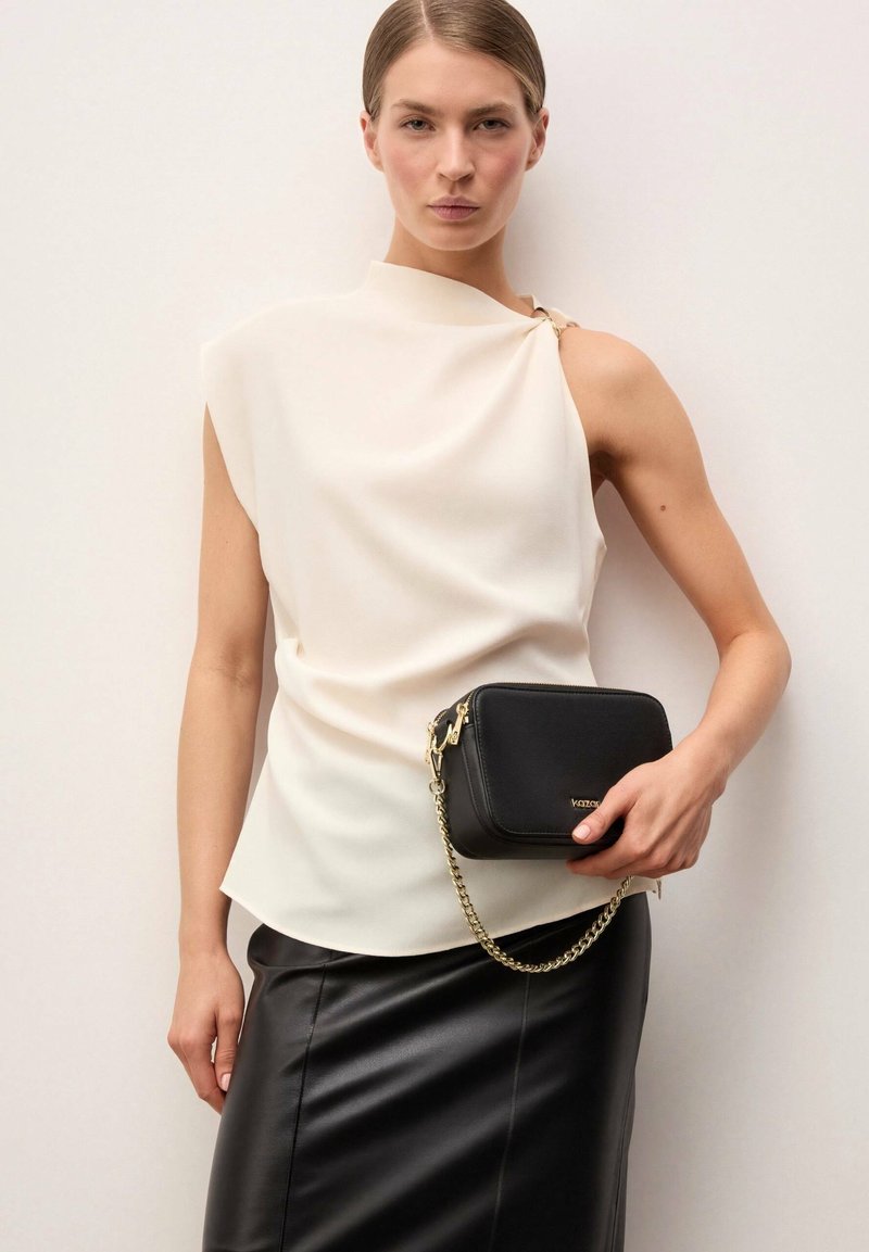 Naine, kes kannab valget varrukateta drapeeritud toppi ja musta nahast seelikut, hoides väikest musta õlakotti kuldse kett-rihmaga.