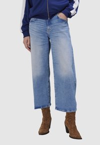 Jean large bleu clair en denim, avec une finition décolorée et un ourlet effiloché, associé à des bottes à cheville à talons en daim marron ornées de motifs.