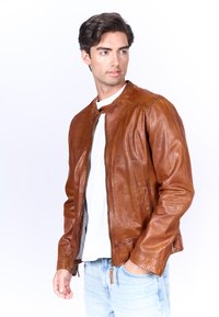 Bruine leren jas met een ronde kraag, ritssluiting aan de voorkant en lange mouwen. Bevat naden en een textuurafwerking. Draagt over een wit shirt.