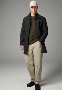 Schwarzer, knöpfbarer Mantel über einem grünen Strickpullover, kombiniert mit beigen Hosen und schwarzen Schuhen. Die Mütze ist hellbeige mit weißer Schrift.