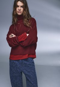 Weinroter übergroßer Pullover mit hohem Kragen, darüber ein rotes gestreiftes Hemd getragen. Strukturierte blaue, gesprenkelte Hose sichtbar.