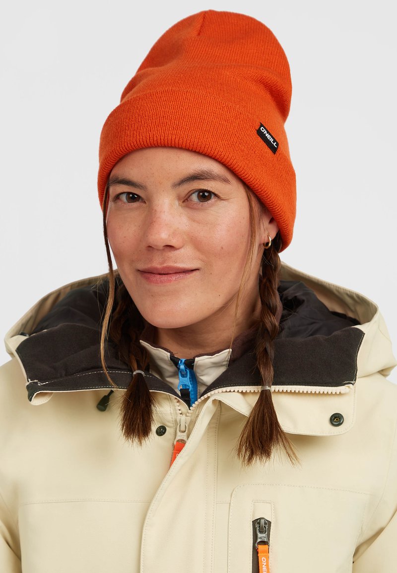 Oranje gebreide muts en een crèmekleurige jas met een donkergrijze kraag. Heeft een blauwe ritssluiting en een oranje accent op de jas.
