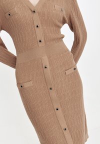 Dzianinowa sukienka w jasnobrązowym kolorze z dekoltem w kształcie litery V, z przodem zapinanym na guziki, dwiema kieszeniami na piersi oraz prążkowaną teksturą na korpusie i rękawach.