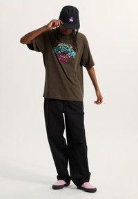 Camiseta verde oliva con un diseño gráfico colorido, pantalones cargo negros y una gorra negra con un logo rosa. Unos zapatos de slip rosa completan el look.