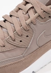 Béžová teniska s texturovanou semišovou částí, síťovanými detaily, silnými tkaničkami a bílou podrážkou. Výraznými prvky jsou vroubkovaná překrytí a logo.