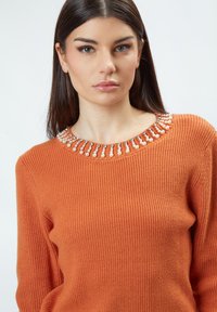Oranje geribde trui met een ronde halslijn, versierd met parelachtige embellishments en kleine strass steentjes rond de kraag.