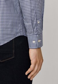 Mano masculina reposando al lado del bolsillo de unos jeans de mezclilla oscuros, vistiendo una camisa de cuadros azul y blanca de manga larga con puños abotonados.