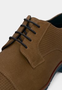 Shoe de vestido em camurça castanha com atacadores pretos, painel lateral texturizado e detalhes na sola em vermelho e azul, visto de um ângulo em close.