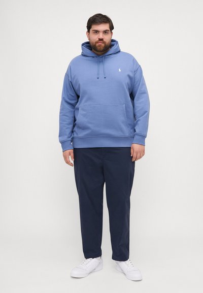 Sudadera de color azul claro con un bolsillo frontal y cordones, combinada con pantalones azul marino y zapatillas blancas. Diseño simple con un logo en el pecho.