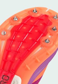 Close-up of a sports shoe sole with orange and red colors, featuring metal spikes for traction, and purple accents on the side.