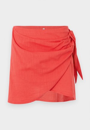 Mini-jupe corail style portefeuille avec un nœud latéral et un tissu subtilement texturé sur un fond clair uni.