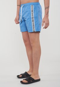 Costumi da bagno azzurri con vita a cordoncino, tasche laterali e strisce nere con logo. Indossati con sandali neri slip-on.