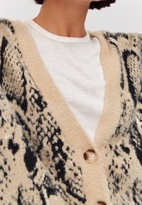 Strickjacke in Beige mit schwarzem Tiermuster. Verfügt über einen V-Ausschnitt, strukturierten Wollmix und Holzknöpfe. Über einem weißen Hemd getragen.