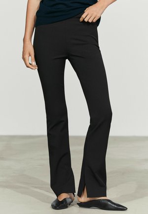 Personne portant un pantalon noir ajusté avec des fentes aux chevilles à l’avant et des chaussures plates pointues noires, debout sur un sol gris clair.