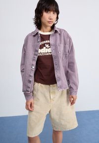 Ljuslila jeansjacka över en brun T-shirt; kombinerad med krämfärgade, knälånga shorts. Modellen poserar mot en enkel bakgrund.