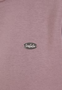 Gros plan d'un tissu tricot mauve avec une petite étiquette ovale en métal argenté cousue, affichant le nom de la marque en cursive.
