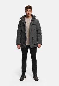 Homme portant un parka d'hiver à capuche gris foncé, un pull beige en maille torsadée, un pantalon noir et des bottes noires, debout devant un fond blanc.