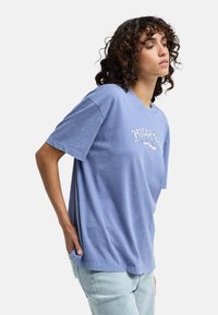 Lichtblauwe katoen t-shirt met korte mouwen, voorzien van een logo-opdruk aan de voorkant. Zachte textuur en een relaxed fit, in een lichte denim jeans gestopt.