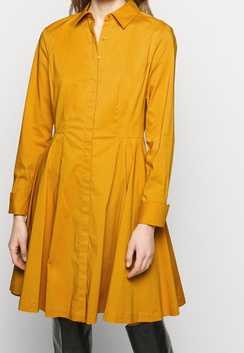 Sukienka koszulowa z długim rękawem w musztardowym kolorze, wykonana z gładkiego materiału. Posiada zapięcie na guziki z przodu, kołnierz oraz rozszerzaną spódnicę z kieszeniami.