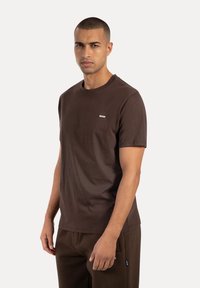 Brązowa bawełniana koszulka z krótkim rękawem, okrągłym dekoltem i małym białym logo na lewej piersi. Gładka tekstura i luźny krój.
