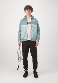 Lyseblå lynlås-træningsjakke med en hvid stribe, båret over en beige T-shirt, parret med sorte joggers og sorte atletiske sko.